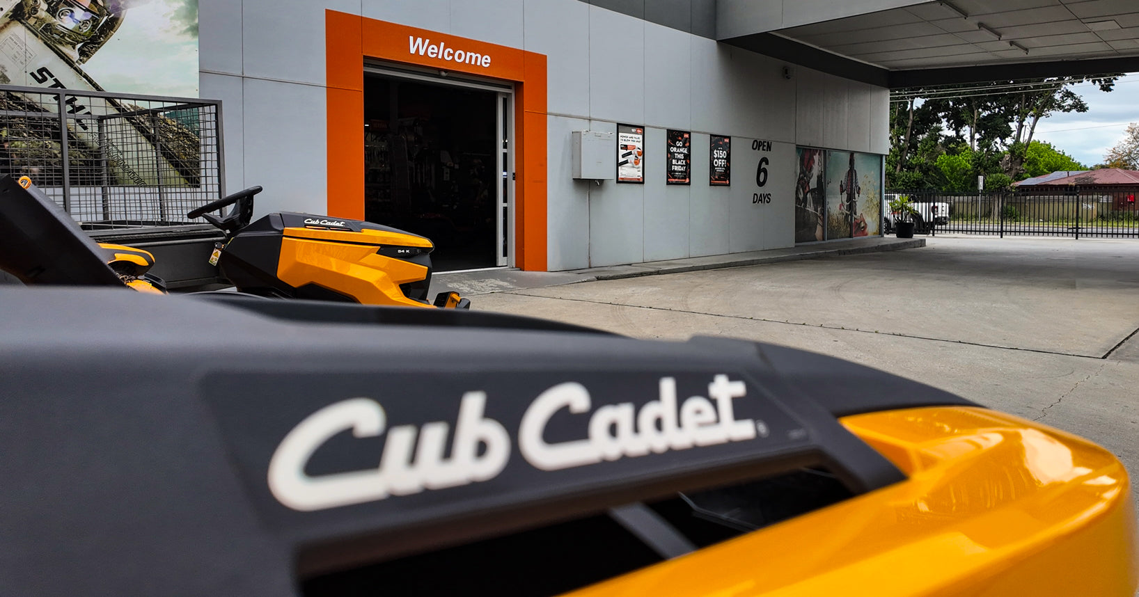 A yellow and black Cub Cadet riding mower in the foreground with the Cub Cadet logo clearly visible on the deck, parked outside Mower City Albury showing the test drive area and welcome sign into the shop