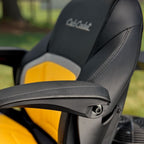 Close-up of a black and yellow Club Cadet outdoor Z3 54 S Zero Turn Mower Chair with a black armrest and the brand name embroidered on the headrest, set against a blurred green background. Talk to Mower City Albury for more details